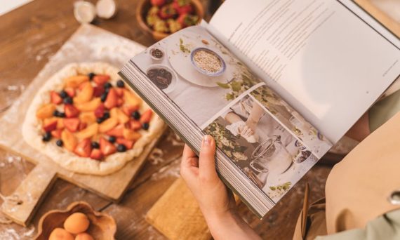 A person reads a cookbook while preparing a fruit tart on a wooden table.